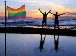 Due persone in riva al mare con le braccia in alto al tramonto, al lato una bandiera rainbow