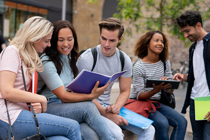 Quattro studenti di diverse nazionalità all'esterno di un campus universitario che parlano e si scambiano appunti, alcuni su tablet e alcuni su quaderno